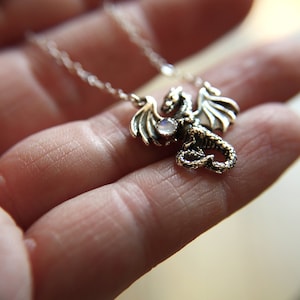 Puede incluir: Un collar con un colgante de dragón plateado adornado con una piedra preciosa transparente. El dragón está detallado con alas y una cola enroscada.