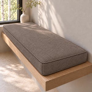 May include: A rectangular bench cushion upholstered in a textured, medium-brown fabric with piped edges. The cushion is positioned on a light-toned wooden bench seat that extends horizontally along a wall near a window.