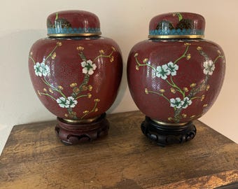 Könnte beinhalten: Zwei passende Cloisonné-Ingwerdosen mit Deckeln, die einen tiefroten Hintergrund mit weißen und grünen Blumenastmotiven aufweisen. Jede Dose hat einen Messingrand und steht auf einem dunklen, geschnitzten Holzständer mit rundem Fuß.
