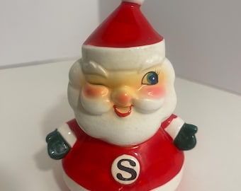 Könnte beinhalten: Eine Keramikfigur des Weihnachtsmanns mit rotem Anzug und weißem Besatz, passender Mütze mit weißem Bommel und dunkelgrünen Fäustlingen. Die Figur hat einen weißen Bart, rote Wangen und zwinkert mit einem Auge. Auf der Brust befindet sich ein schwarzes S.