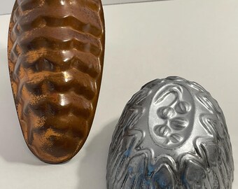 Könnte beinhalten: Zwei Backformen aus Metall. Eine ist eine längliche, kupferfarbene Form mit einem strukturierten, sich wiederholenden geometrischen Muster. Die andere ist eine silberfarbene, kuppelförmige Form mit einem erhabenen Reliefmuster aus kleinen Beeren auf der Oberseite.