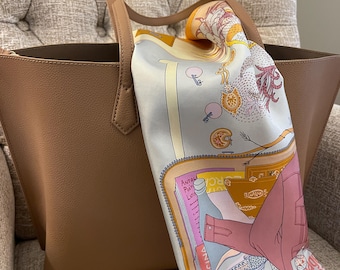 Könnte beinhalten: Eine beigefarbene Ledertasche mit zwei Schulterriemen, über die ein großes, rechteckiges Seidentuch drapiert ist. Das Tuch zeigt einen farbenfrohen Grafikdruck mit verschiedenen Objekten, darunter eine rosa Jacke, Schuhe und der Text "magic car".