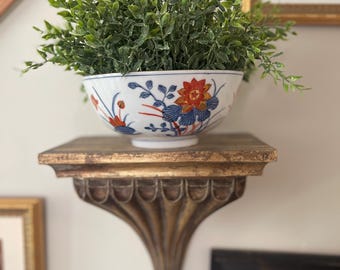 Könnte beinhalten: Eine weiße Keramikschale mit einem handbemalten Blumenmuster in Rot-, Blau- und Goldtönen. Die Schale ist mit einer üppigen grünen Kunstpflanze mit kleinen, dichten Blättern gefüllt, die einen lebendigen Kontrast zum weißen Porzellan bildet.