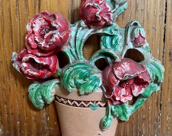Könnte beinhalten: Eine Wandplakette aus Gussmetall mit einem beigefarbenen Blumentopf, der am Rand ein Zickzackmuster aufweist und mit geformten roten Mohnblumen und grünen Blättern gefüllt ist. Das Stück hat ein abgenutztes Finish mit sichtbarem Farbabrieb.