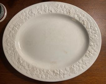 May include: An oval white ceramic serving platter featuring a raised, embossed floral and fruit border pattern around the rim. The center of the platter is smooth and plain, with a glossy finish that reflects light across its surface.