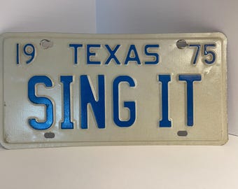 Puede incluir: Una placa de matrícula rectangular de metal blanco con letras y bordes azules en relieve. La placa presenta el texto 19 TEXAS 75 en la parte superior y la palabra SING IT en grandes caracteres azules en el centro, con cuatro orificios de montaje.