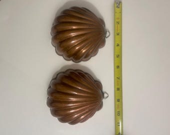 Könnte beinhalten: Zwei kupferfarbene, muschelförmige Formen mit gerippter Textur und kleinen Metallaufhängungen. Ein gelbes Maßband liegt neben den Formen und zeigt, dass jede Muschel eine Länge von etwa 12,7 cm aufweist.