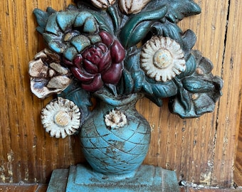 Könnte beinhalten: Ein Türstopper aus Gussmetall mit einem Blumenstrauß in einer blauen Vase. Das Arrangement besteht aus weißen, roten und bläulichen Blüten mit grünen Blättern. Die Vase hat ein Rautenmuster und steht auf einem rechteckigen Sockel mit Patina.