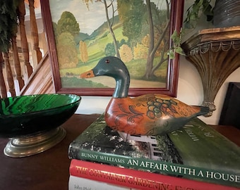 Könnte beinhalten: Eine dekorative Enten-Lockvogel-Figur aus Holz mit einem dunkelgrünen Kopf, einem roten Auge, einem gelben Schnabel und einem orangefarbenen Körper, der mit handgemalten schwarzen Schnörkeln und grünen Trauben verziert ist, auf zwei Büchern.