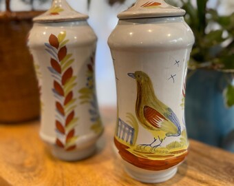Könnte beinhalten: Zwei cremeweiße Keramikdosen mit passenden Deckeln, jeweils mit handgemalten Motiven. Eine Dose zeigt einen gelben Vogel mit roten und blauen Flügeldetails, während die andere ein vertikales Muster aus roten, gelben und blauen Blättern aufweist.