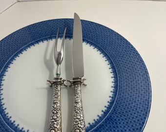 Könnte beinhalten: Ein silberfarbenes Tranchierbesteck bestehend aus Gabel und Messer mit detaillierten floralen Prägegriffen und Parierstange, platziert auf einem weißen Teller mit breitem blauem Rand und Punkt- sowie Bogenmuster.