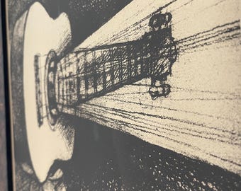 May include: A framed black and white ink illustration featuring a guitar positioned on a stand. The artwork uses high-contrast cross-hatching techniques to depict the instrument, with a light-colored beam highlighting the neck and headstock.