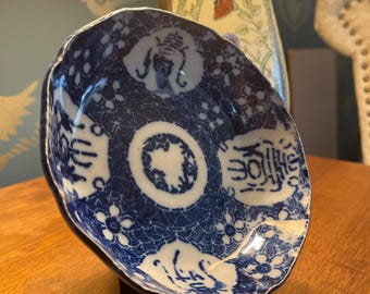 Könnte beinhalten: Eine kleine, runde Keramikschale mit gewelltem Rand und blau-weißem Muster. Das Design umfasst komplexe geometrische Wellenmotive, florale Akzente und vier markante kreisförmige Felder, die jeweils stilisierte chinesische Schriftzeichen enthalten.