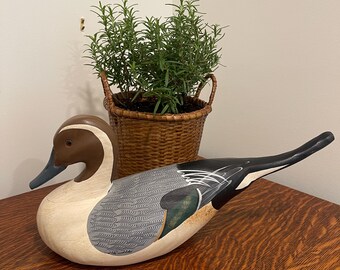 Könnte beinhalten: Eine handbemalte Enten-Lockvogel-Figur aus Holz mit braunem Kopf und weißem Streifen, grauem Körper mit detaillierter Federzeichnung, dunkelgrünem Flügelbereich und einem langen, spitz zulaufenden schwarzen Schwanz in ruhender Haltung.