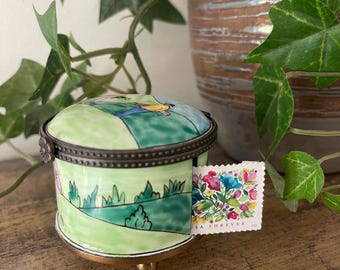 Könnte beinhalten: Eine runde Schmuckdose aus Porzellan mit einer handgemalten grünen Landschaftsszene und einer Figur im gelben Hemd. Der Deckel hat ein Metallscharnier und eine florale Briefmarke mit der Aufschrift USA FOREVER steckt an der Seite.