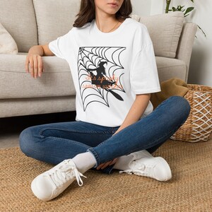 May include: A white short-sleeved t-shirt featuring a black graphic of a spider web and a witch silhouette. The text "It's spooky season witches" is printed in orange across the centre of the design. The shirt has a crew neckline and a relaxed fit.