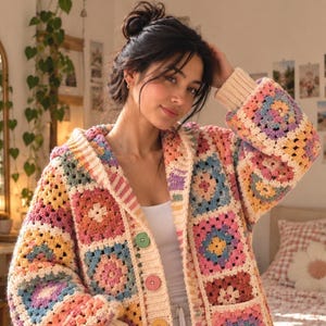 Peut inclure: Un cardigan en crochet à manches longues avec un motif de carrés granny colorés dans des tons de rose, jaune, bleu et violet. Le vêtement comprend une capuche, des poignets côtelés et une fermeture boutonnée avec trois boutons ronds vert, rose et jaune.