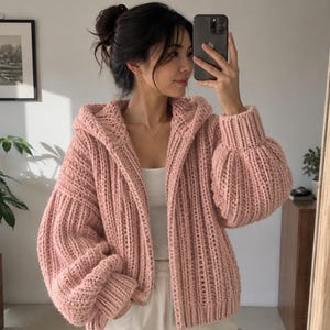 May include: A chunky knit cardigan in a soft pastel pink hue, featuring a hood, long voluminous sleeves with ribbed cuffs, and a vertical textured stitch pattern. The garment is worn open over a simple white top, showcasing its thick, cosy construction.