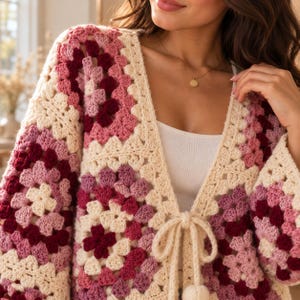 May include: A long-sleeved crochet cardigan featuring a multi-colored granny square pattern in shades of cream, light pink, mauve, and deep burgundy. The garment has a V-neckline and a front tie closure made of cream-colored yarn with pom-pom ends.