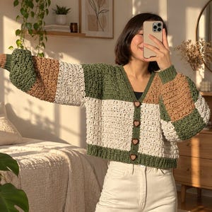 Peut inclure: Un cardigan en crochet à manches longues avec un motif color-block en vert olive, crème et beige. Le vêtement présente une encolure en V, des poignets et un ourlet côtelés, et se ferme sur le devant par quatre boutons marron en forme de cœur.