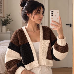 May include: A color-blocked crochet cardigan featuring horizontal stripes in dark brown, black, and cream. The garment has a V-neckline, long sleeves with ribbed cuffs, and a ribbed hem. The design is constructed with a textured stitch pattern.