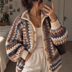 Peut inclure: Un gilet en crochet avec un motif granny square dans des tons bleu, marron, crème et beige. Le vêtement présente un col, des manches longues avec des poignets côtelés et une fermeture boutonnée sur le devant avec des boutons ronds marron clair.