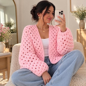 Peut inclure: Un cardigan rose clair ouvert sur le devant, doté d'un motif au crochet en carrés de grand-mère avec de longues manches volumineuses. Le vêtement est porté sur un débardeur blanc simple et un jean bleu clair, offrant un aspect tricoté texturé.