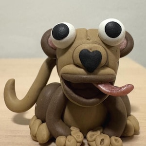May include: A sculpted clay monkey figurine featuring a light brown body, dark brown limbs, and large white eyes with black pupils. The monkey has a black heart-shaped nose, a wide open mouth, and a pink tongue sticking out to the side.