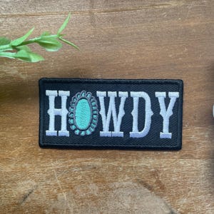 May include: A rectangular black embroidered patch featuring the white text HOWDY, with the letter O replaced by a turquoise oval stone design surrounded by a dark grey scalloped border. The patch has a black stitched edge and a flat rectangular shape.