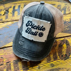 May include: Distressed gray baseball cap with a tan mesh back. A white patch on the front reads "Pickle Ball" in black script with a pickleball graphic. The cap has a pre-worn look with distressed details.