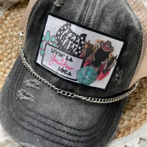 May include: A black distressed trucker hat with a mesh back and a silver chain. The hat has a patch with a colorful design and the text "Livin' La Yea Haw Loca".