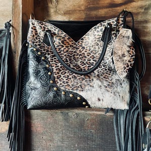 Puede incluir: Un bolso de mano negro y con estampado de leopardo con asa de cuero y flecos. El bolso presenta una combinación de cuero negro repujado, estampado de leopardo y piel de vaca. Tachuelas doradas acentúan el cuero negro. Un charm de pluma de cuero a juego cuelga del asa.