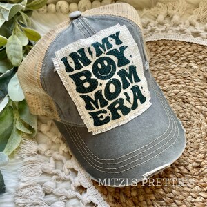 May include: Gray trucker hat with a distressed brim and a tan mesh back. The hat features a patch with the text "IN MY BOY MOM ERA" in dark green, with a smiley face and lightning bolt accents.