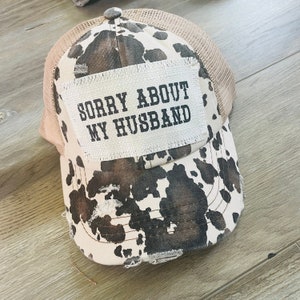 May include: A distressed baseball cap with a cow print design in brown and white. The cap features a mesh back and a patch with the text "SORRY ABOUT MY HUSBAND" in black.