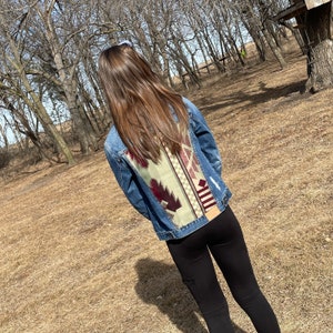 May include: A denim jacket with a decorative back panel featuring a geometric pattern in shades of green, burgundy, and cream. The jacket has a classic design with distressed details.