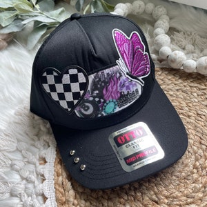 May include: Black baseball cap with a checkered heart, a purple butterfly, and a multi-colored patch with a pool ball. The cap has a classic fit and a mid profile. The label reads "OTTO PREMIUM FABRIC CLASSIC FIT ONE SIZE MID PROFILE MAXIMUM COMFORT"