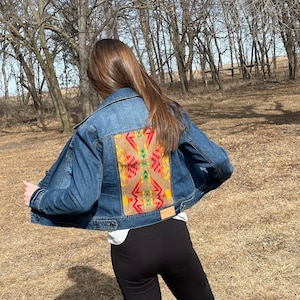 May include: A blue denim jacket with a colorful geometric pattern on the back. The pattern is in shades of red, yellow, green, and brown.
