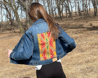 Puede incluir: Una chaqueta vaquera azul con un patrón geométrico colorido en la espalda. El patrón está en tonos de rojo, amarillo, verde y marrón.