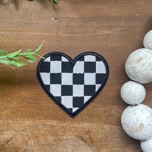 Peut inclure: Un écusson brodé en forme de cœur avec un motif à damier noir et blanc. L'écusson présente une bordure cousue en fil noir qui souligne la forme du cœur, créant un contraste marqué entre les carrés sombres et clairs du motif géométrique.