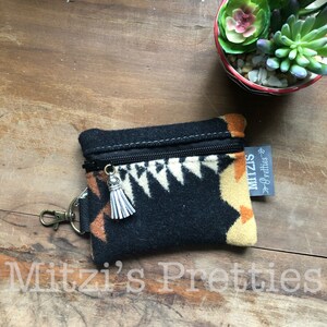 May include: A small, black zippered pouch with a geometric pattern in cream and orange. It has a silver tassel zipper pull and a metal clasp. The text "Mitzi's Pretties" is visible on a tag.