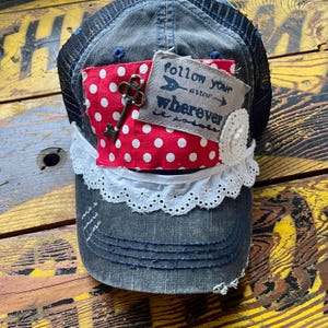 May include: A denim trucker hat with a black mesh back, adorned with a red polka dot patch, a vintage key, and a fabric tag that reads "follow your arrow wherever it points". The hat features white lace trim.