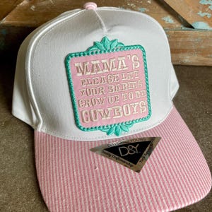 May include: A white baseball cap with a pink and white pinstriped brim and a pink button on top. A rectangular patch features the embroidered text MAMA'S PLEASE LET YOUR BABIES GROW UP TO BE COWBOYS in white, framed by a teal decorative border.