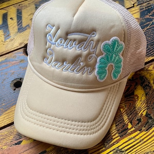 May include: Tan trucker hat with a mesh back and a curved brim. The hat features the embroidered text "Howdy Darlin" in white script, along with a green cactus patch. The hat is designed with a classic trucker style.