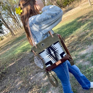 May include: A green canvas tote bag with a black and white geometric pattern. The bag has a brown leather trim and a long strap. The bag is being worn by a person wearing a light blue denim jacket and blue jeans.