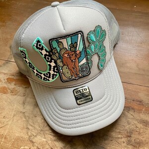 May include: A light gray trucker hat with a mesh back and foam front. The front features a black and white patterned horseshoe patch, a rectangular patch with a brown cow and cactus illustration, and a turquoise decorative accent patch. OTTO sticker.