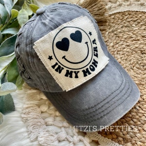 May include: Gray baseball cap with a distressed finish. The cap features a square patch with a smiley face design, heart-shaped eyes, and the text "IN MY MOM ERA". The cap has a braided detail on the back.
