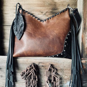 Puede incluir: Bolso de hombro de cuero marrón con flecos negros y hoja decorativa. El bolso presenta costuras negras y tachuelas plateadas. El bolso tiene una correa negra y está sobre un fondo de madera.