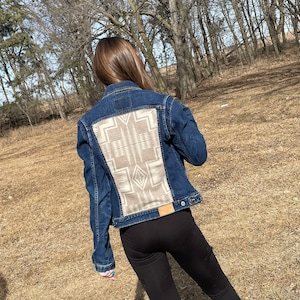 May include: A blue denim jacket with a tan and white patterned fabric panel on the back. The fabric panel features a geometric design.