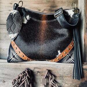 Puede incluir: Bolso de hombro de piel de vaca con un diseño en blanco, negro y marrón. El bolso tiene una correa de cuero, flecos decorativos y cierre de cremallera. Está adornado con tachuelas metálicas.