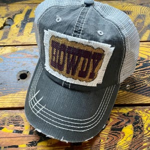 May include: A distressed charcoal gray baseball cap with a white mesh back. The front features a rectangular patch with the word "HOWDY" in brown letters on a burlap-like background. White stitching accents the brim.